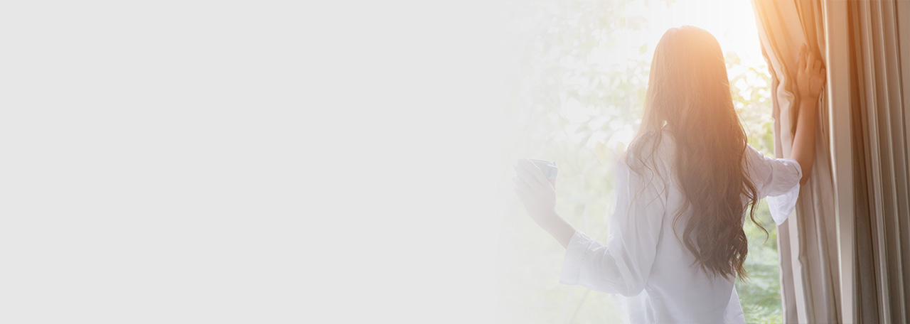 Nainen katsoo ulos pidellen verhoja.